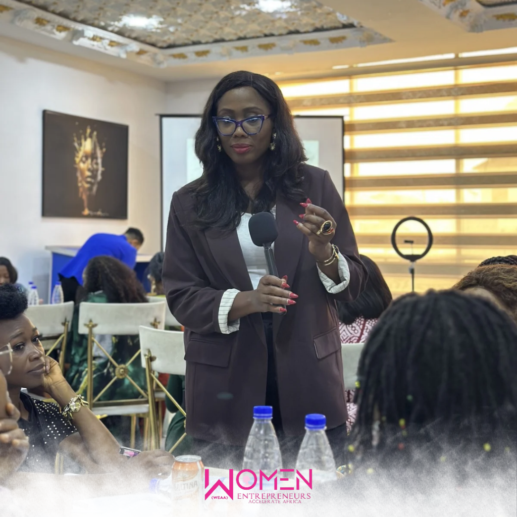 Image of a woman standing with a microphone and speaking in the midst of other seated women at a conference