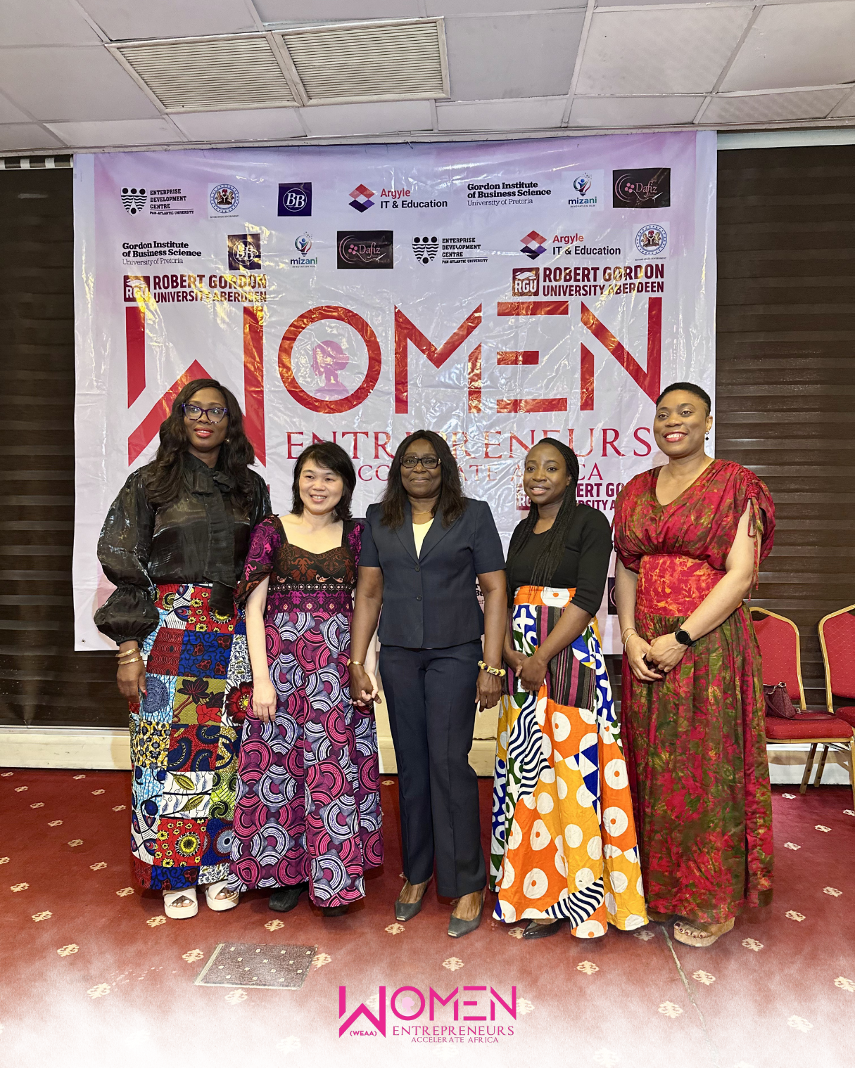 5 women standing in front of a banner from an event and smiling to the camera