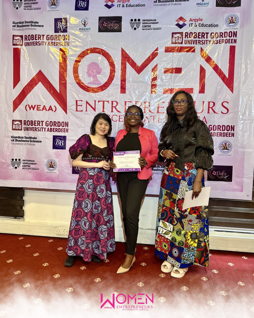 3 women standing in front of an event banner that boldly reads 'WOMEN WEAA ENTREPRENEURS' The one in the middle holding a certificate and smiling
