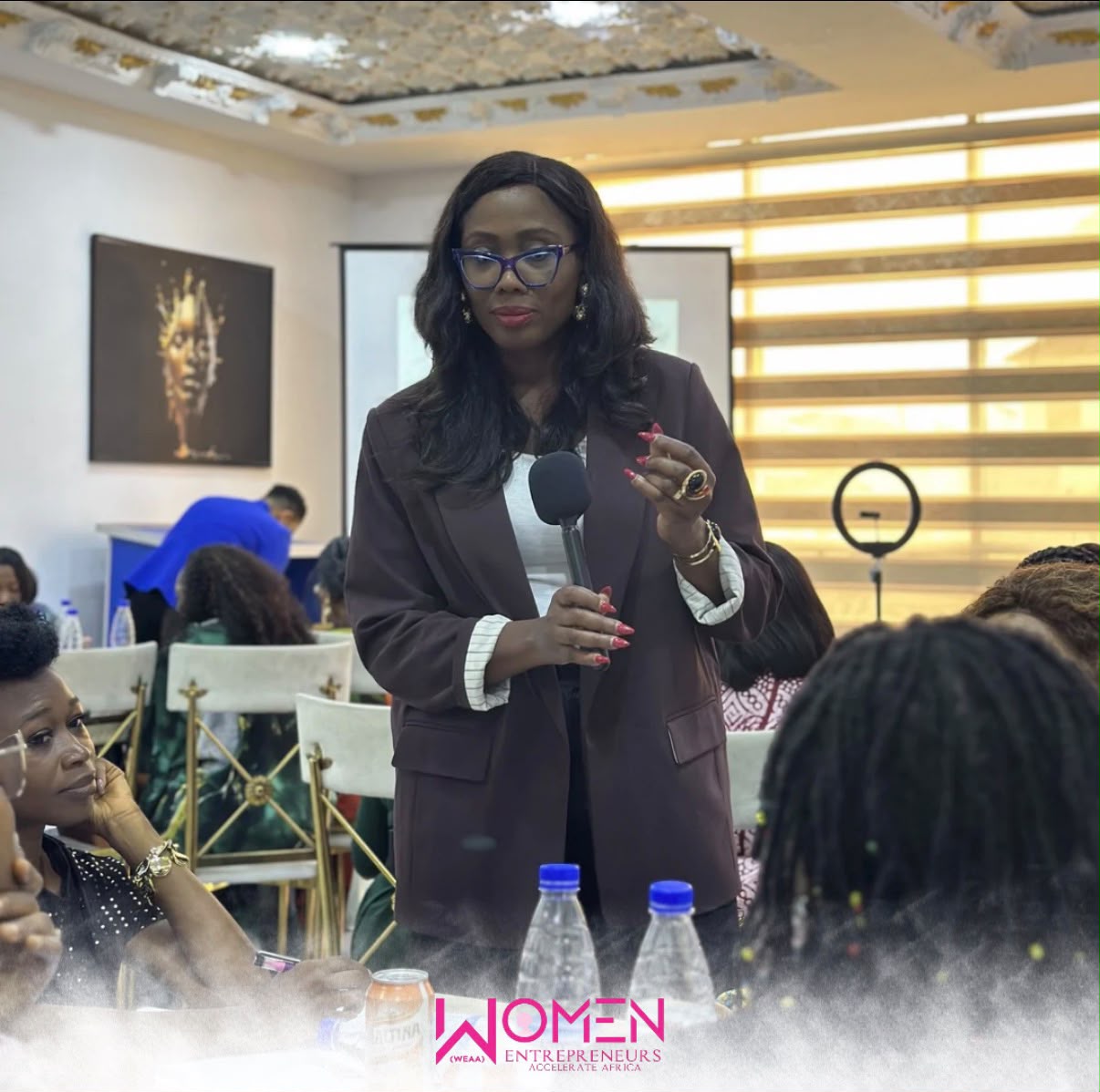 Image of a woman standing with a microphone and speaking in the midst of other seated women at a conference