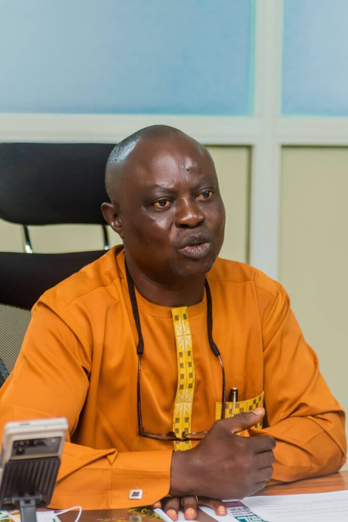 A black African male wearing an orange outfit, speaking while seated in an office facing the right side of the photo