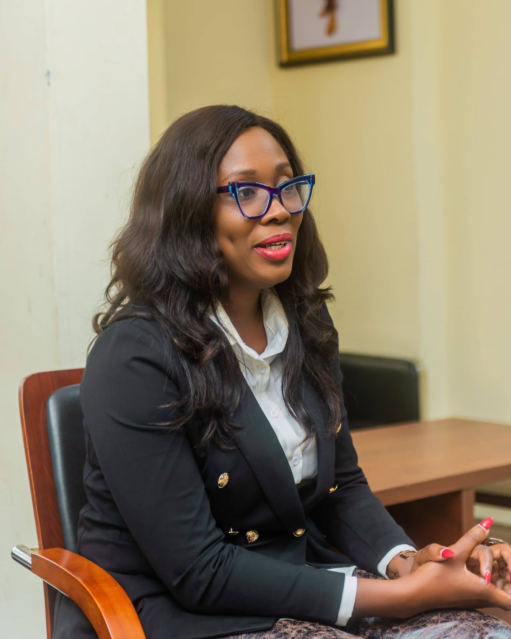 A black African female wearing a black jacket and glasses seated in an office facing the right side of the photo