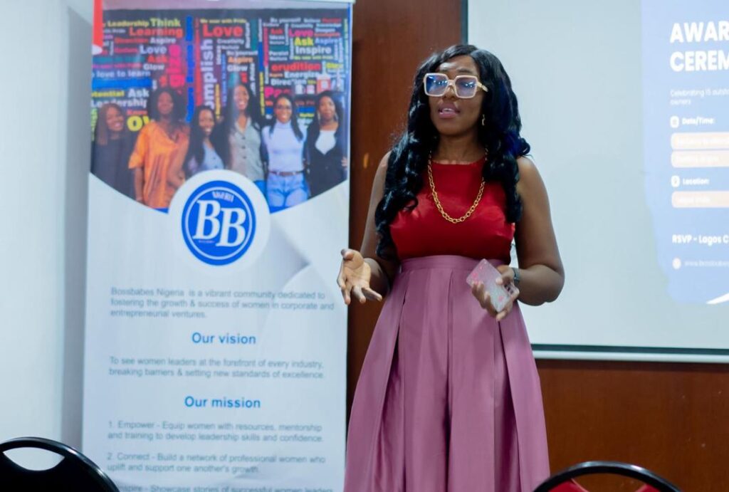 A lady wearing a red top and glasses, standing in front of an event banner and speaking.